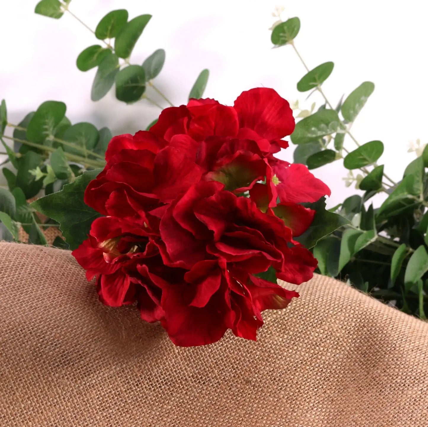 Vibrant red artificial flowers with green eucalyptus leaves on burlap, perfect for home décor or gift arrangements