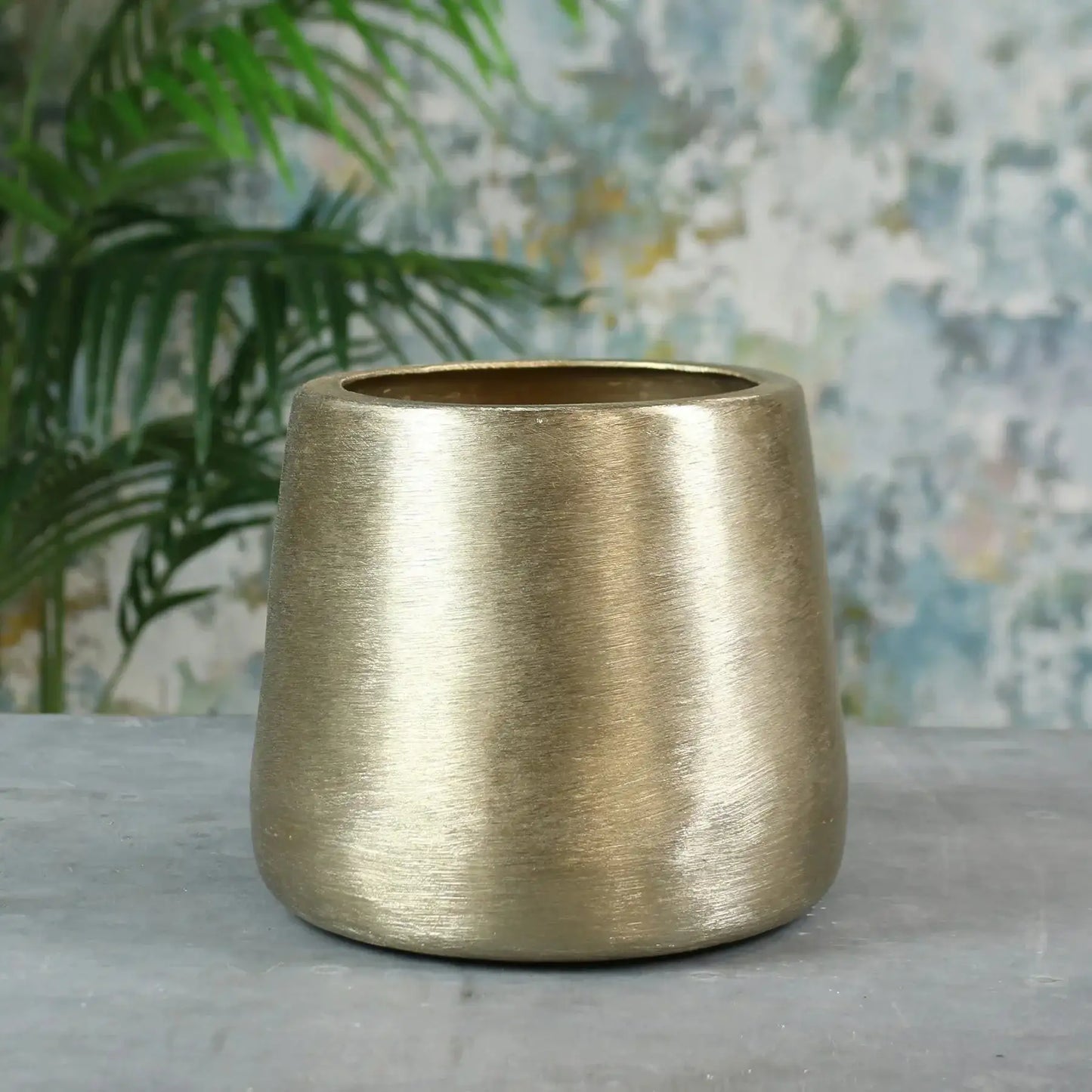 Brushed gold metal planter pot on a grey surface with leafy green plant in background