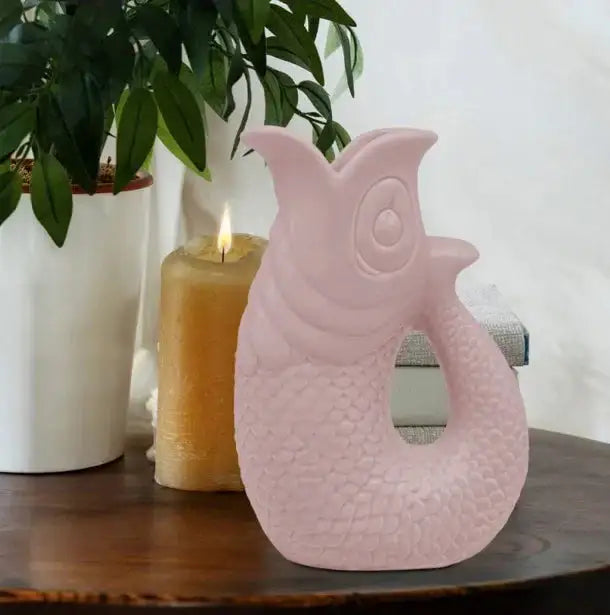Pink fish-shaped decorative vase on wooden table with lit candle and potted plant