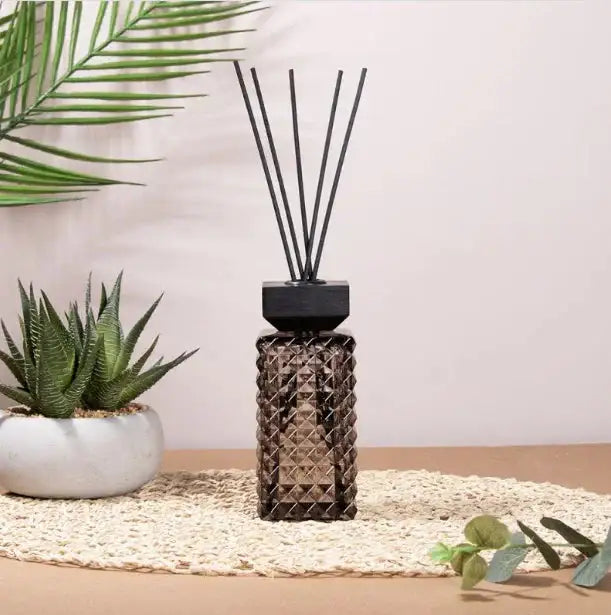 Diamond-patterned reed diffuser with black sticks on a mat, with a potted succulent and palm leaf decor.
