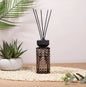 Diamond-patterned reed diffuser with black sticks on a mat, with a potted succulent and palm leaf decor.
