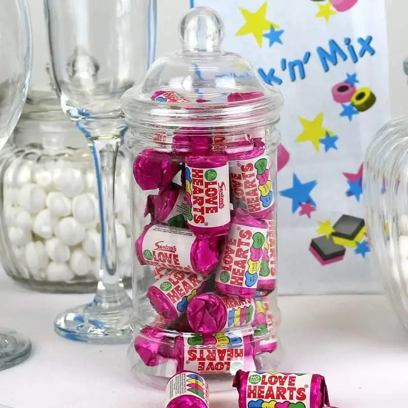Glass jar filled with Swizzels Love Hearts candies, surrounded by glassware and sweets.